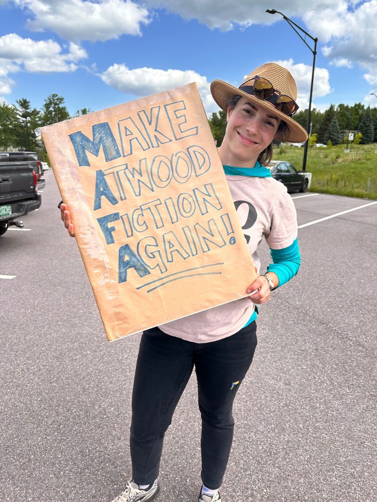 A woman in a wide brimmed hat standing holding a sign that says Make Atwood Fiction Again.