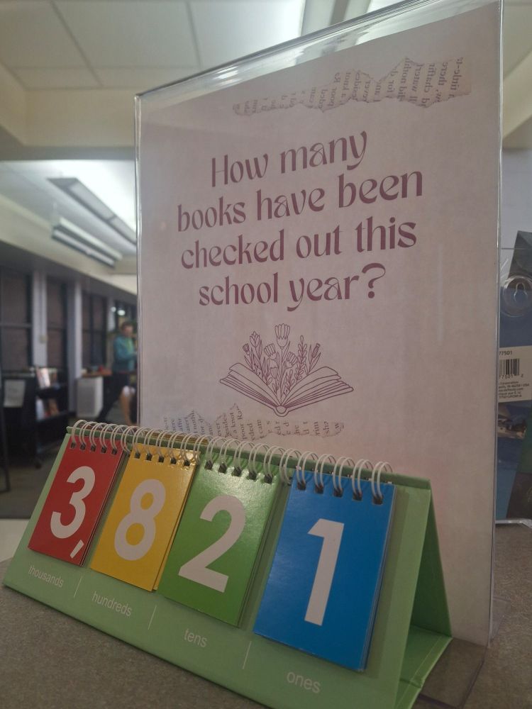 A sign on the library desk that says "how many books have been checked out this school year?" In front of it is a flip chart that currently shows the number 3,821. 