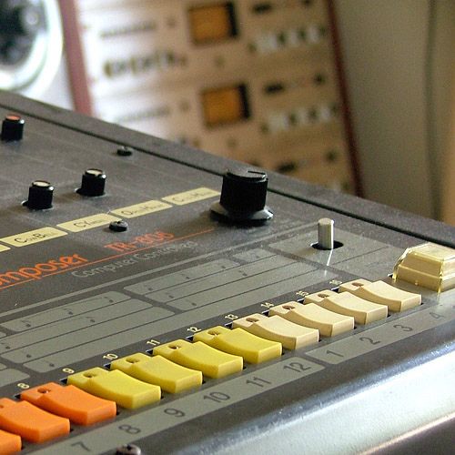 A photo of a TR-808.  Alas, I don't know who took it.  It's a fun, very early '80s looking drum machine, with its iconic row of red, orange, yellow, and white buttons against a black background.