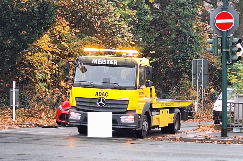 Ein gelber LKW mit dem Aufdruck "Meister" und "ADAC" steht mit eingeschalteter oranger Warnblinkanlage an einer Ampel. Rechts von ihm ist ein Teil eines roten Kleinwagens zu erkennen. 