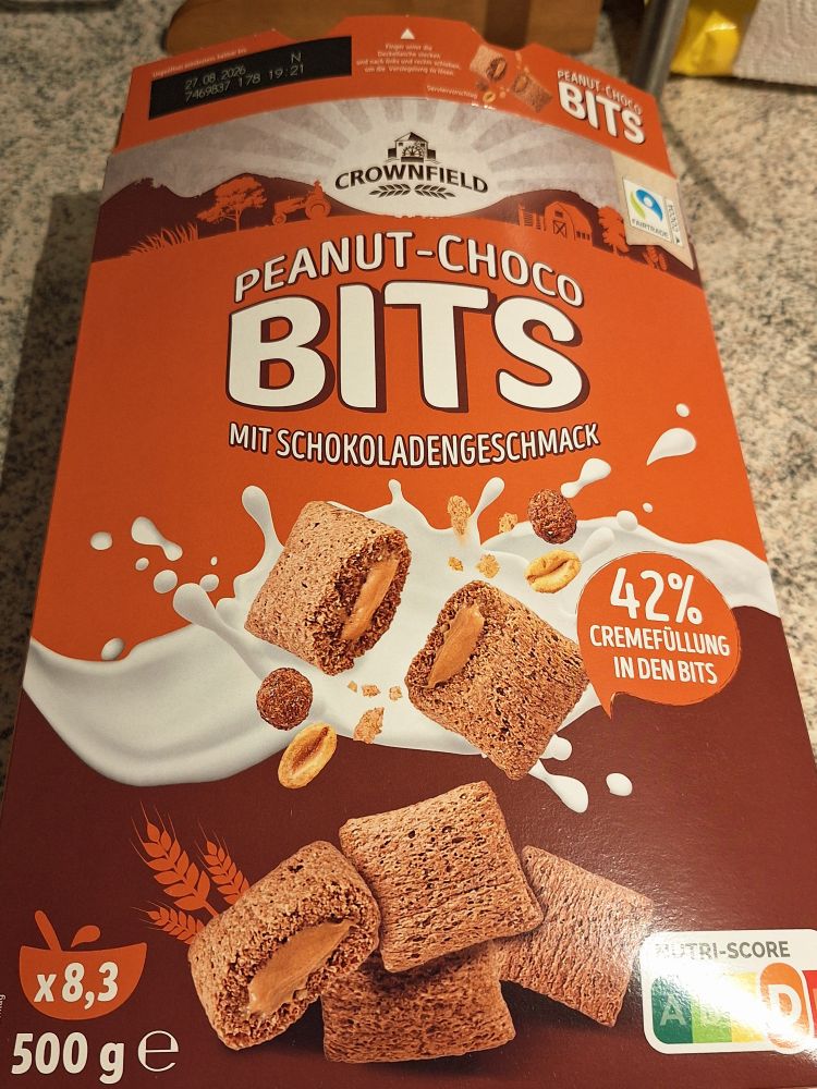 Eine orange und braun gefärbte Cornflakes-Packung mit dem Schriftzug Crownfield Peanut-Choco Bits mit Schokoladengeschmack, 42% Cremefüllung in den Bits, 500g entsprechen 8,3 Portionen. Ws sind auseinandergebrochene braune Knusperkissen mit erdnussbutterfarbener Füllung aufgedruckt.