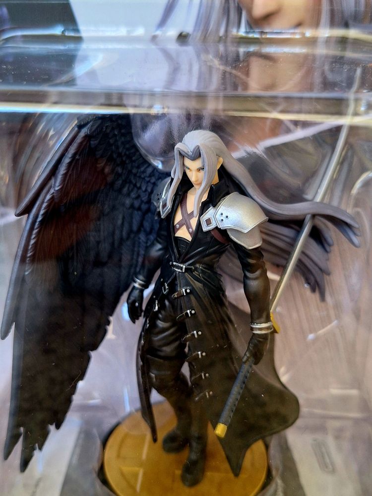 Nahaufnahme des Amiibos durch die Packung. Sephiroth steht in seiner schwarzen Hose und dem langen schwarzen Mantel mit den silbernen Schulterstücken da. In der linken Hand hält er sein sehr langes schmales Schwert Masamune. Seine silbernen langen Haare flattern im Wind. Er hat rechts einen großen Flügel mit schwarzen Federn.