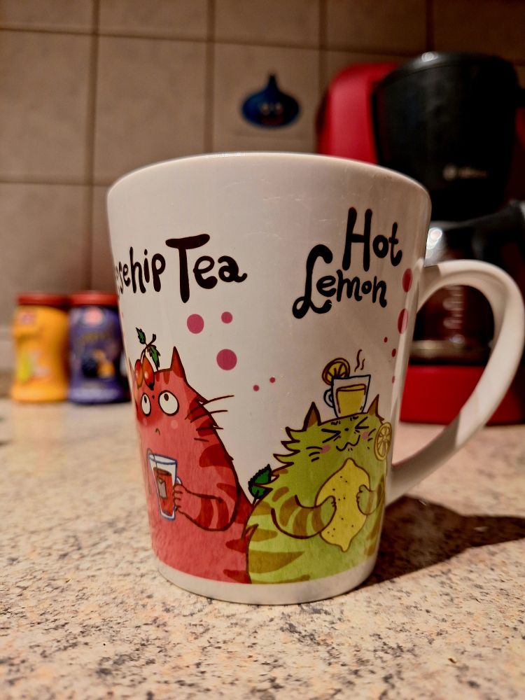 Eine weiße Tasse auf der zwei Comic-Katzen zu sehen sind. Eine pink-rote mit einem Glas rotem Tee mit Teebeutel drin in den Pfötchen und einer Hagebutte auf dem Kopf, zu der sie hoch starrt. Und eine grüngelbe Katze mit verkniffenem Gesicht, einer Zitrone in den Pfötchen und einem Glas heißer Zitrone auf dem Kopf.