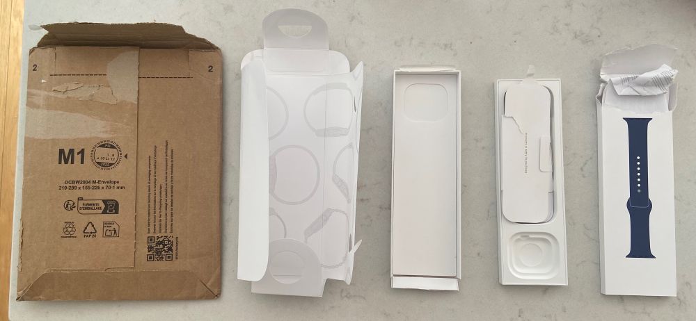 Various bits of packaging laid out left to right with the Amazon envelope far left and empty torn Apple Watch packaging towards the right. 