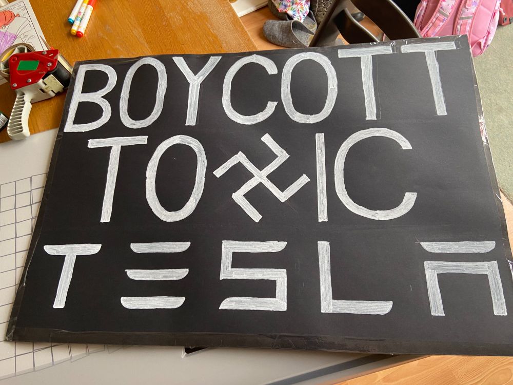 A protest sign with white text on black background and the text BOYCOTT TO卍IC TESLA