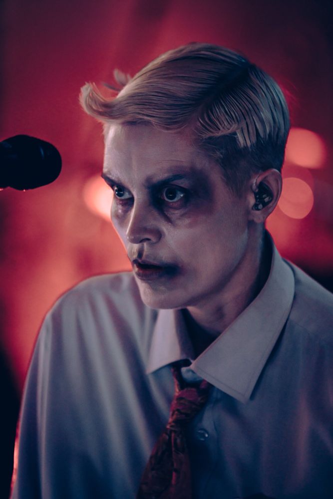 Karin Dreijer du projet Fever Ray arborant un maquillage dramatique avec un teint pâle et des cernes sombres, évoquant une ambiance gothique ou surnaturelle. Les cheveux sont coiffés avec précision dans un style rétro, et l'artiste porte une chemise blanche avec une cravate rouge légèrement dénouée. L'éclairage rougeâtre et tamisé crée une atmosphère intense et énigmatique, avec un micro visible à gauche, suggérant une performance musicale ou théâtrale.