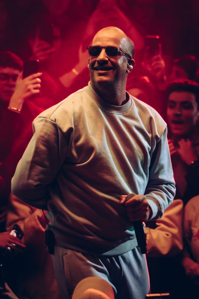 RIMK souriant en pleine performance, vêtu d’un ensemble de survêtement clair. Il porte des lunettes de soleil rondes et tient un micro dans sa main droite. L'éclairage rouge intense ajoute une atmosphère énergique à la scène, tandis que le public en arrière-plan est capturé en train de profiter du moment, certains levant leurs téléphones pour immortaliser l’instant.