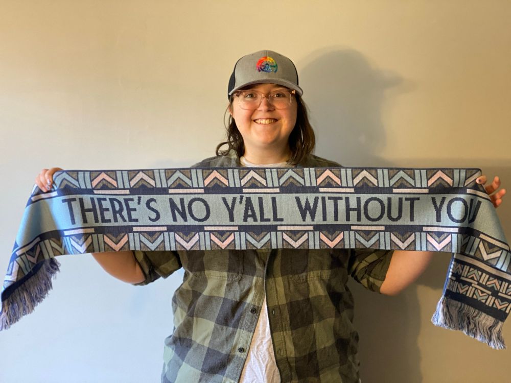 Brittany in a grey hat with a rainbow Asheville City logo, holding a scarf she designed for the South Slope Blues, with art deco designs inspired by Asheville and text “There’s no y’all without you”