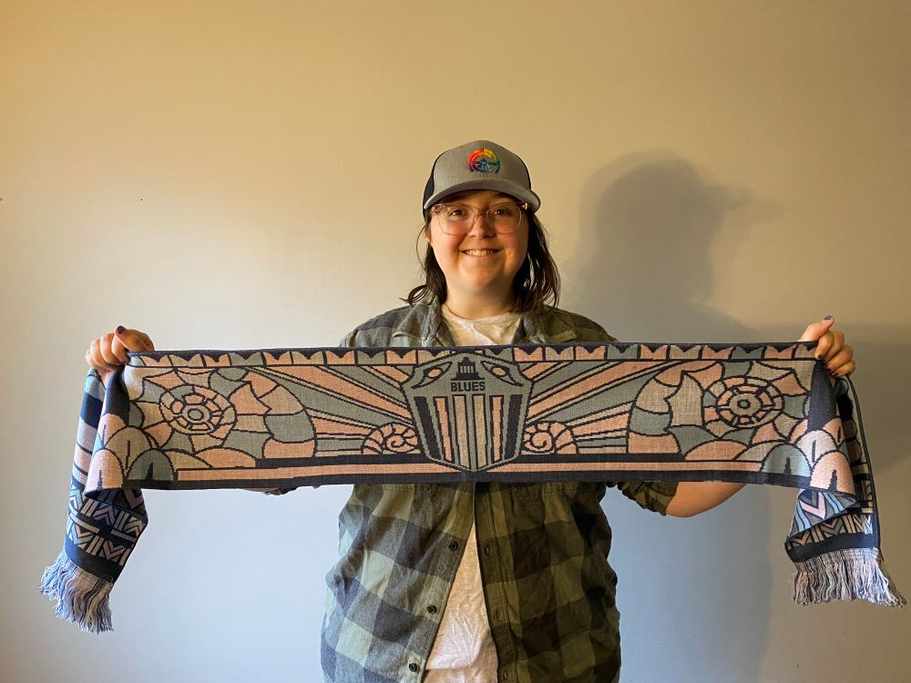 Brittany in a grey hat with a rainbow Asheville City logo, holding a scarf she designed for the South Slope Blues, with art deco designs inspired by Asheville