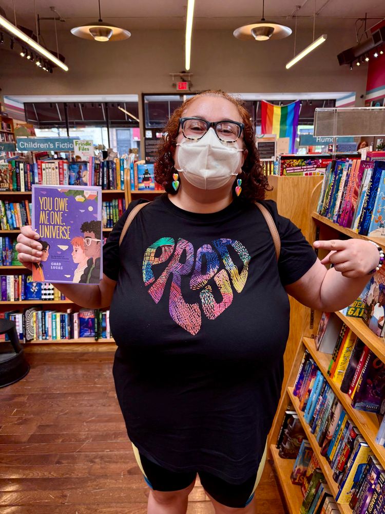 Daniele grins gleefully in a bookstore, wearing her Pride outfit and a white face mask. She points to a copy of the book “You Owe Me One, Universe,” a middle grade contemporary novel by her client Chad Lucas. End alt text. 