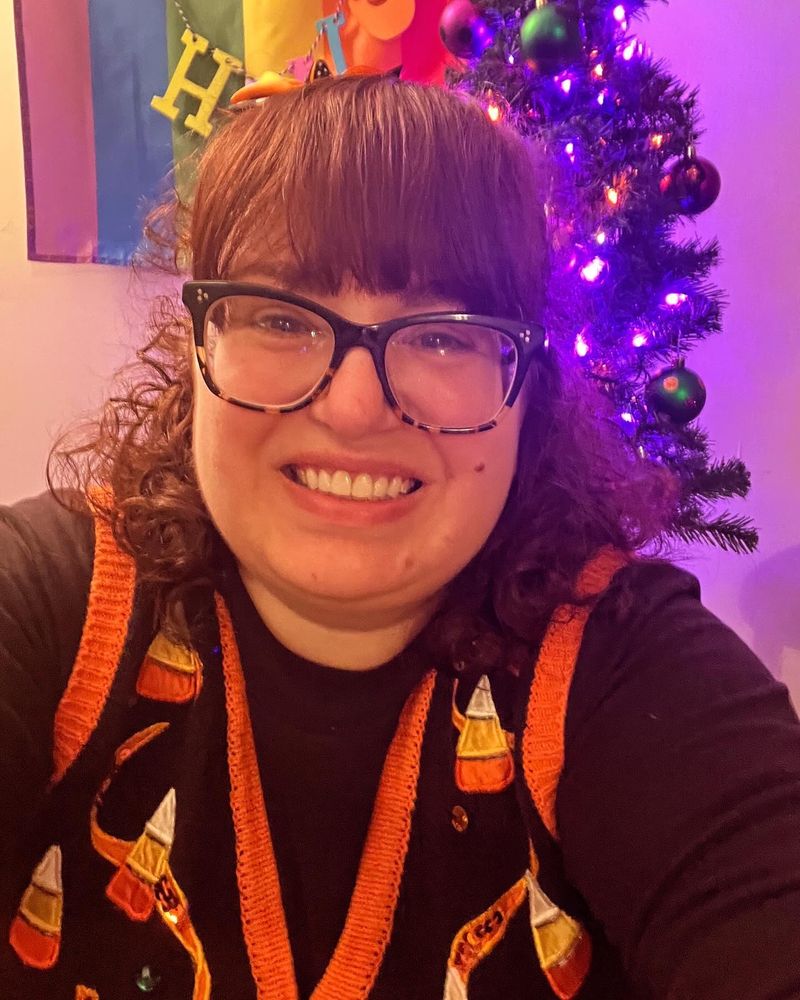 Daniele is a woman with curly auburn hair, straight bangs, and glasses. She wears a sequined vest with candy corns and jack-o-lanterns, smiling at the camera. A small tree with purple and orange lights can be seen behind her. End alt text.