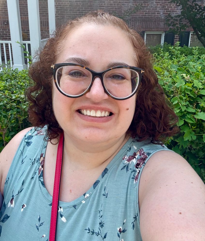 Daniele is a white woman with curly auburn hair and large cat-eye glasses. She smiles, wearing a teal floral dress. End alt text. 
