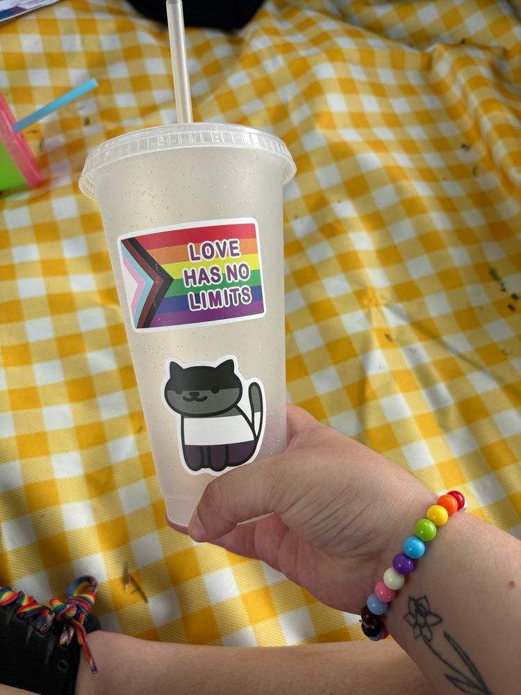 Daniele’s white arm with a daffodil tattoo holds up a clear sparkly cup. It’s decorated with two stickers: The first is a Progress Pride flag with “love has no limits” written on it, and an illustrated cat in the colors of the asexual flag. End alt text. 