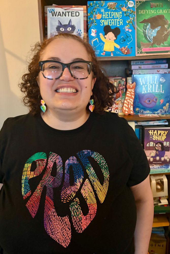 Daniele is a short, fat white woman with curly auburn hair and glasses. She wears a black shirt with the word “proud” written in the shape of a heart, in rainbow textured letters. She grins in front of her bookshelf. End alt text. 