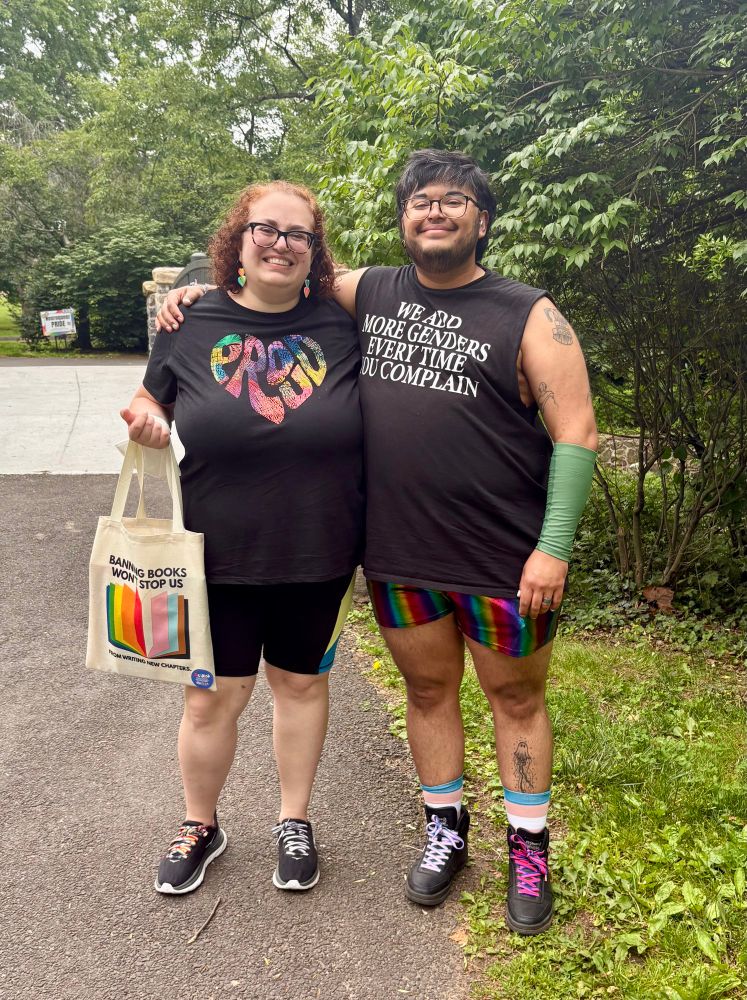 Daniele stands in her Pride outfit holding a tote bag that reads “banning books won’t stop us from writing new chapters.” Her arm is around her husband Quincy, a Brown trans guy with black hair, beard, and glasses. His black tank top reads, “We add more genders every time you complain.” End alt text. 