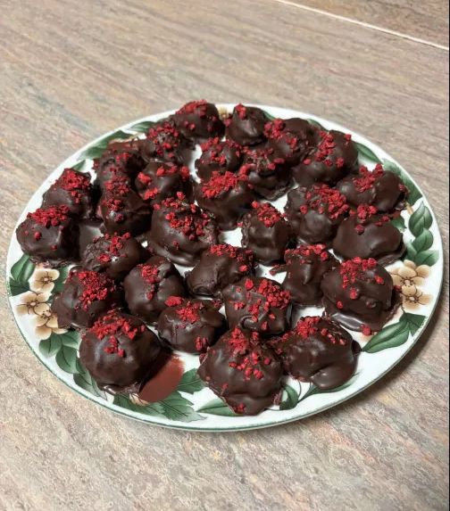 A plate of a little under 30 dark chocolate raspberry truffles.  There are bits of crushed freeze-dried raspberry sprinkled on top. 