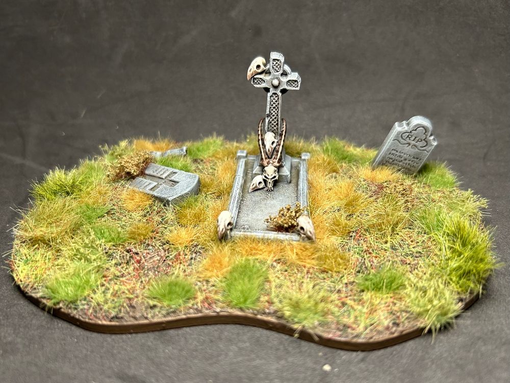 A small piece of wargaming scatter terrain. Three graves two of which are broken and in disrepair. The central one is well maintained, a large flat table tomb with a Celtic cross. Scattered over the grave are bird skulls and a large deer skull rests against the base of the cross. Various tufts make the ground look unkempt and wild.