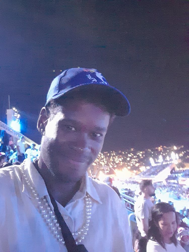 Homem negro sorri para foto. Veste boné azul, colar de pérolas e uma pochete atrevassada. Ao fundo, a Sapucaí 
