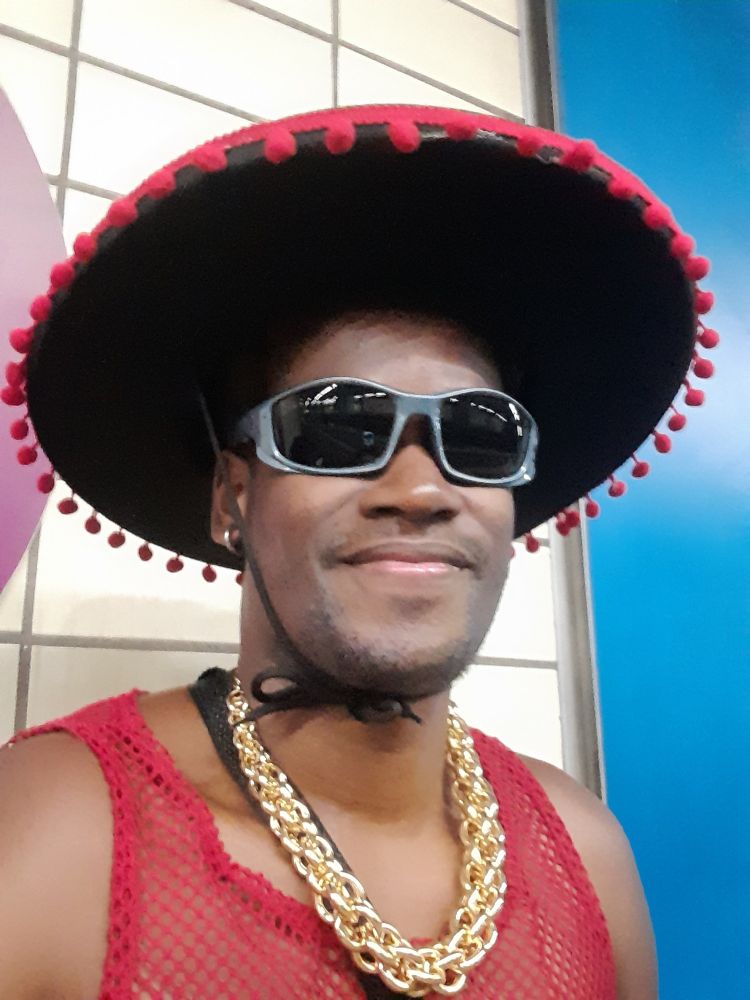 Homem negro veste chapéu preto, cordão dourado, brincos, óculos e uma regata de tela. Olha pra câmera e sorri. Fantasiado para o Carnaval