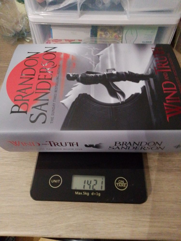 A thick hardcover book with a greyscale cover with red highlights showing a man in formal fantasy clothing standing in front of a mountain and a huge red moon on a kitchen scales. Scales read 1421 grams. The book is Wind and Truth, Stormlight Archive Book Five, by Brandon Sanderson
