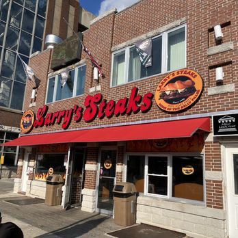 The image shows the exterior of a Philadelphia restaurant named Larry’s Steaks. The building features a red awning with the restaurant’s name prominently displayed in bold red letters. The logo includes an image of a cheesesteak sandwich, and the entrance is flanked by large windows and trash bins. Flags and banners are visible above the awning, and the brick façade gives the establishment a casual appearance.