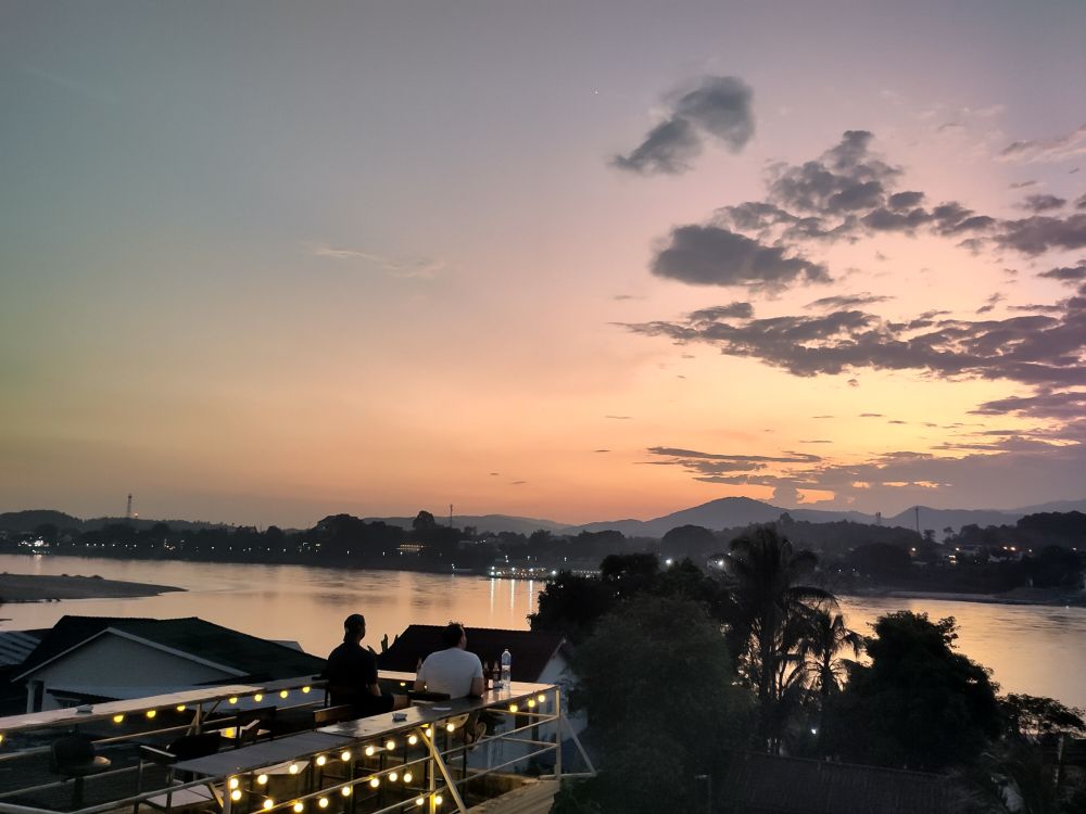 Im Hintergrund ein Sonnenuntergang hinter Bergen. In der Mitte der Mekong. Vorne eine Dachterasse mit 2 Personen.