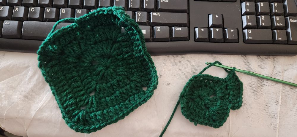 An image of a finished granny square and one in the works, both using an emerald yarn. The yarn works are laid on white tablecloth with a keyboard in frame behind them.