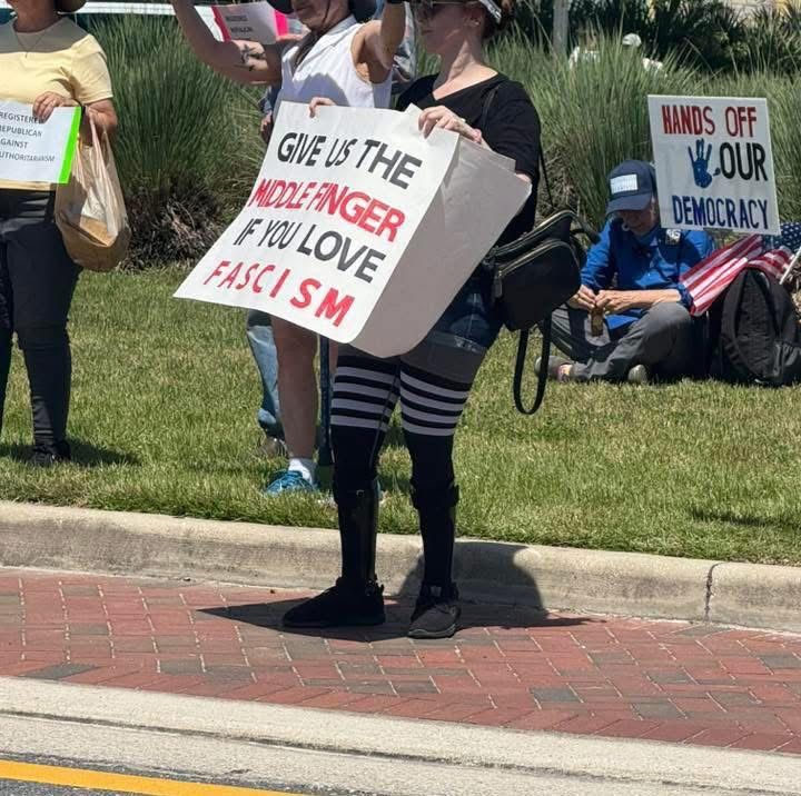 Person holding sign that reads “Give Us The Middle Finger If You Love Fascism”.