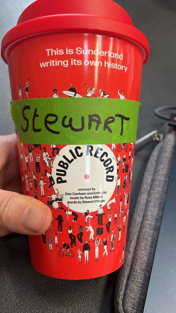 Red keep-cup with the artwork for the community theatre show Public Record and a green name tag reading ‘Stewart’