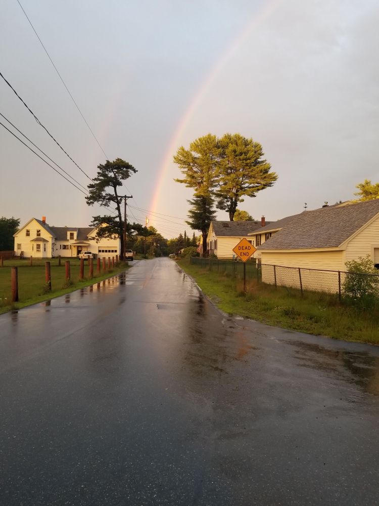 Photo looking down a street with a dead end sign. A rainbow touches down seemingly at the end of the street. 