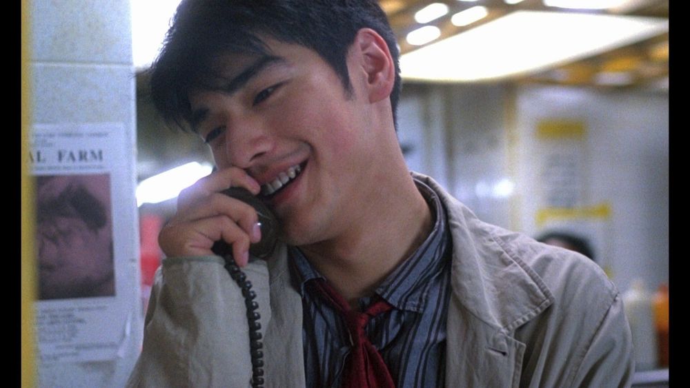 Actor Takeshi Kaneshiro, shown in the film Chungking Express (directed by Wong Kar-Wai), wears a tan jacket over a bllue shirt as he smiles and talks into a black telephone