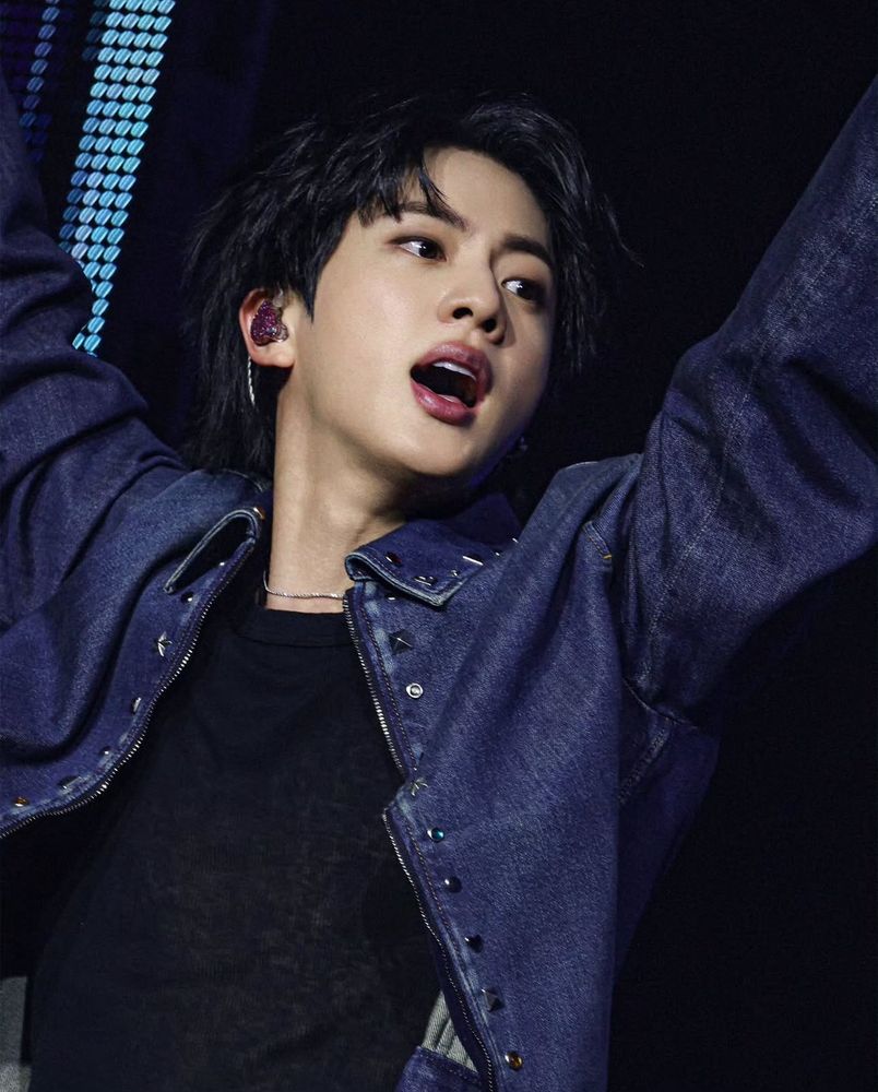 Seokjin in denim jacket over black almost sheer top, with his arms raised