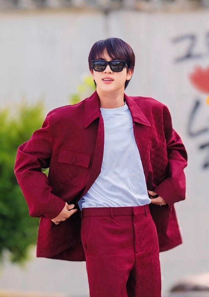 Seokjin wearing a burgundy suit and black sunglasses standing confidently outdoors. He is slightly pulling open his jacket to reveal a  white shirt underneath.