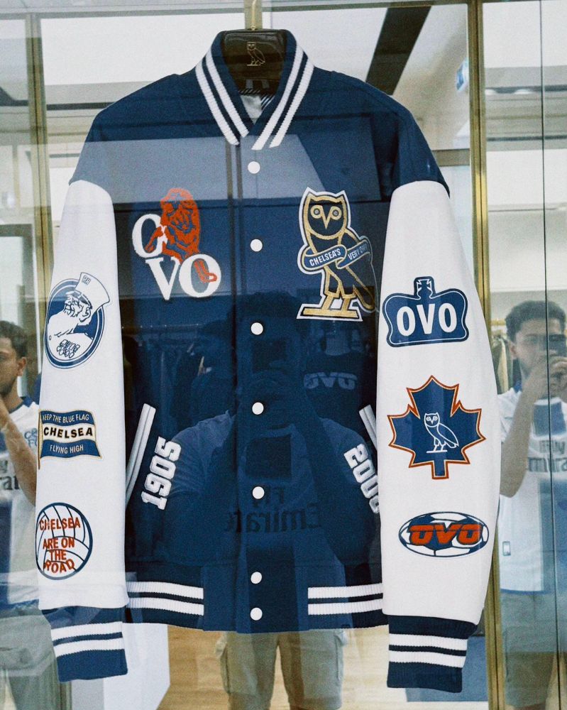 A navy blue and white varsity jacket, a collaboration between Chelsea FC and Drake's OVO brand, is displayed in a glass case. The jacket features numerous patches:
 * Chest: A red Chelsea lion over "CFC" and "OVO" on the left, and the OVO owl logo within a crest on the right.
 * Sleeves: The white sleeves are adorned with various patches, including the "OVO" text logo, a Canadian maple leaf with the OVO owl, and circular patches with Chelsea slogans like "KEEP THE BLUE FLAG FLYING HIGH".
 * Body: The years "1905" (Chelsea's founding year) and "2005" are embroidered vertically in white on either side of the snap buttons.
The reflection of the person taking the photo with their phone is visible in the glass.