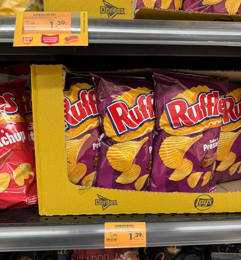 A supermarket shelf displaying a cardboard tray of Ruffles jamon-flavoured potato chips in purple bags. Other varieties of chips, including Lay's, are on the shelves above and below. A price tag shows €1.39.