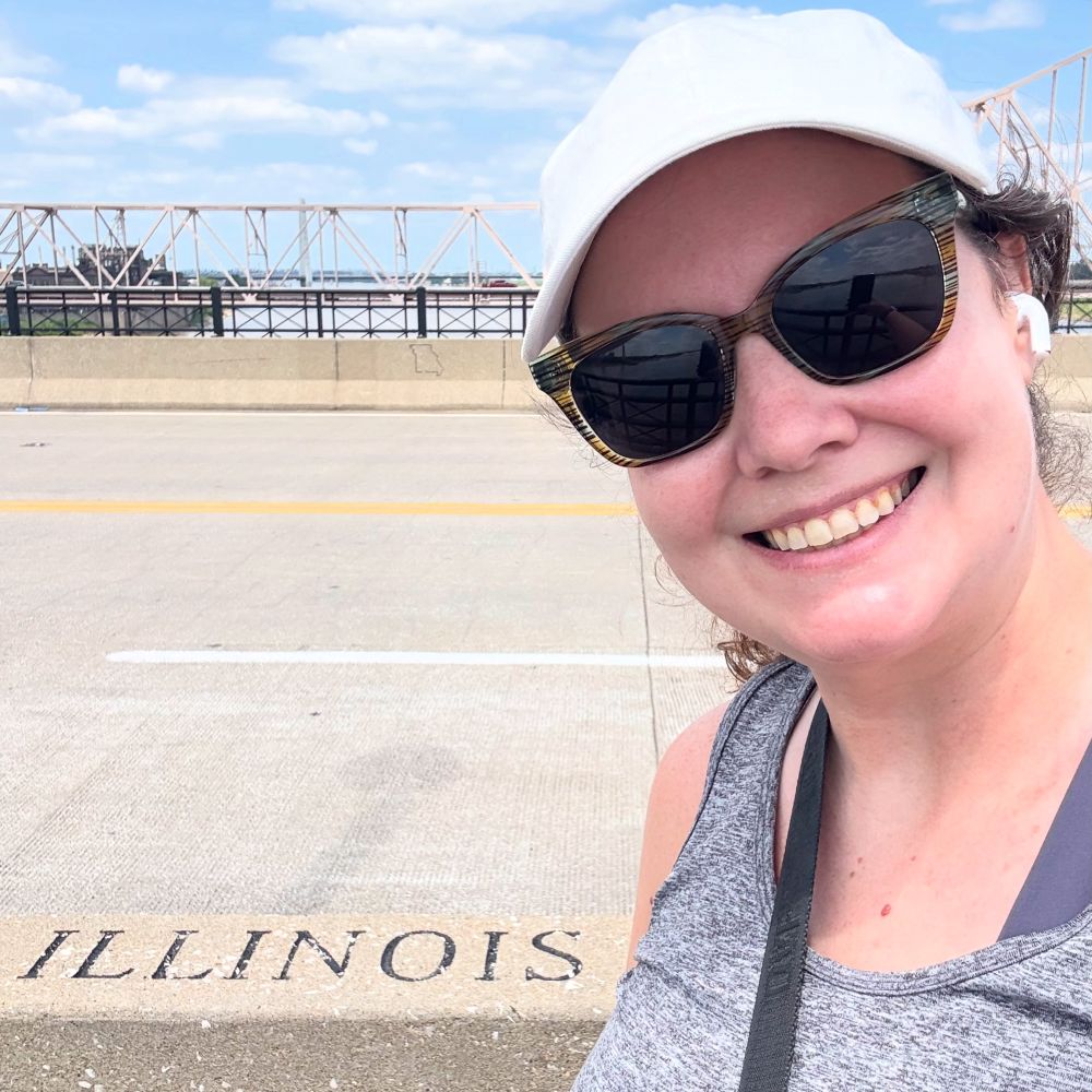A photo of me, a middle aged white woman wearing workout clothes including a gray tank, white hat, and sunglasses, on the Eads Bridge. The photo is a selfie and behind me is the word Illinois, as I’ve just crossed the state line. 