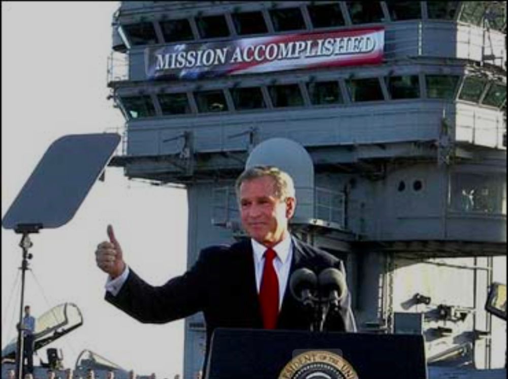 George Bush standing behind a lectern on the flight deck of an aircraft carrier. Giving a thumbs up behind him is a banner that says mission accomplished