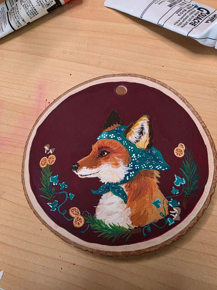 Small painting on a wood slice of a fox wearing a green bandana, on a wine red background. Surrounded by a frame of ivy, pine, mushrooms, and orange slices.