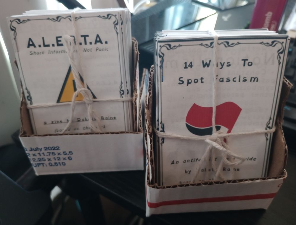 Two bundles of pocket-size zines in stands; "A.L.E.R.T.A." and "14 Ways To Spot Fascism".