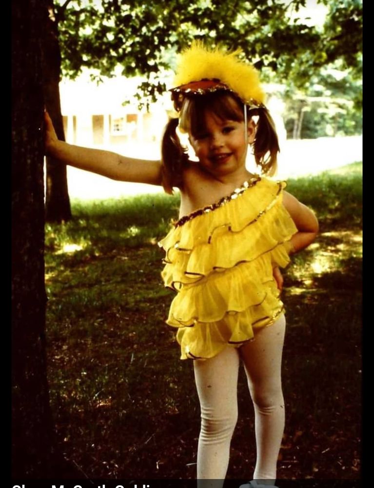 A white child with blonde hair around three years old with pigtails wearing a layered yellow dance suit meant to look like a duck. 