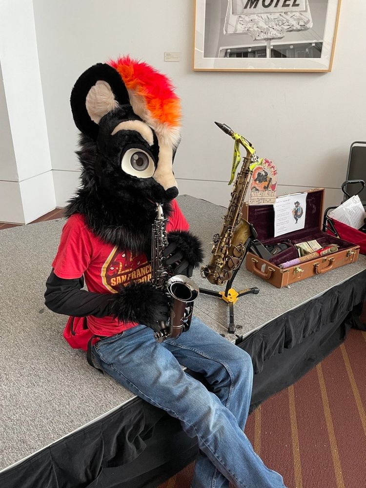 Tech sitting down, playing his Soprano Saxophone, Raven. Dressed in a red San Francisco Hydra t-shirt and jeans. In the background is his Alto Saxophone, Dogfish, and a case for busking tips.