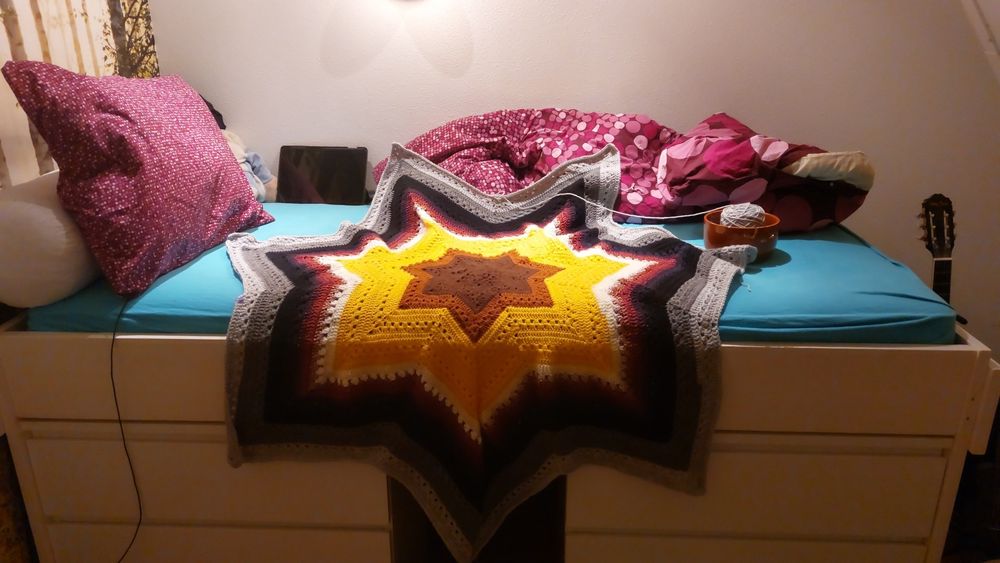 An unfinished crochet 7-point-star blanket, made with (from inside to outside) brown, yellow, white, crimson, black, dark grey and light grey yarn. It is draped over the side of a high bed.