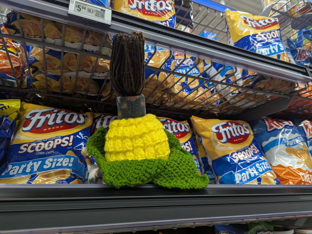 A photograph of a cornservant plushie among bags of corn chips