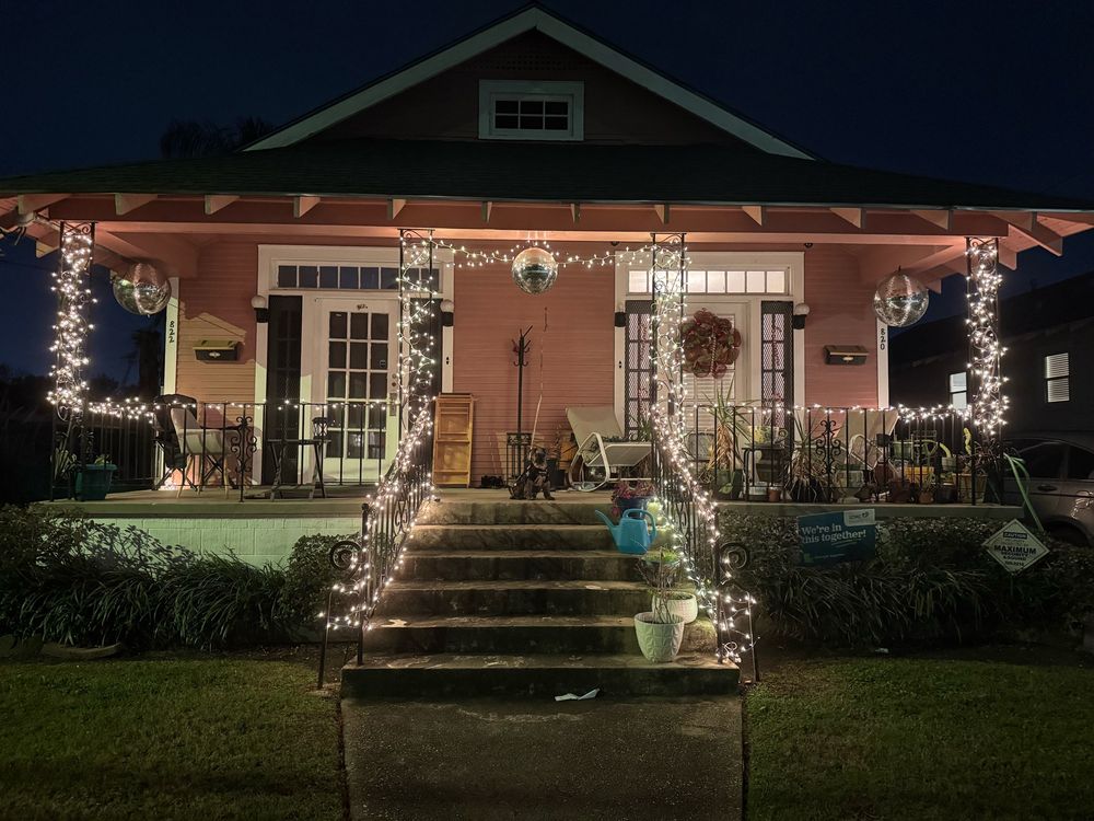 A pink double shotgun house trimmed in white Christmas lights and disco balls, with a small schnauzer sitting in the center very proudly 
