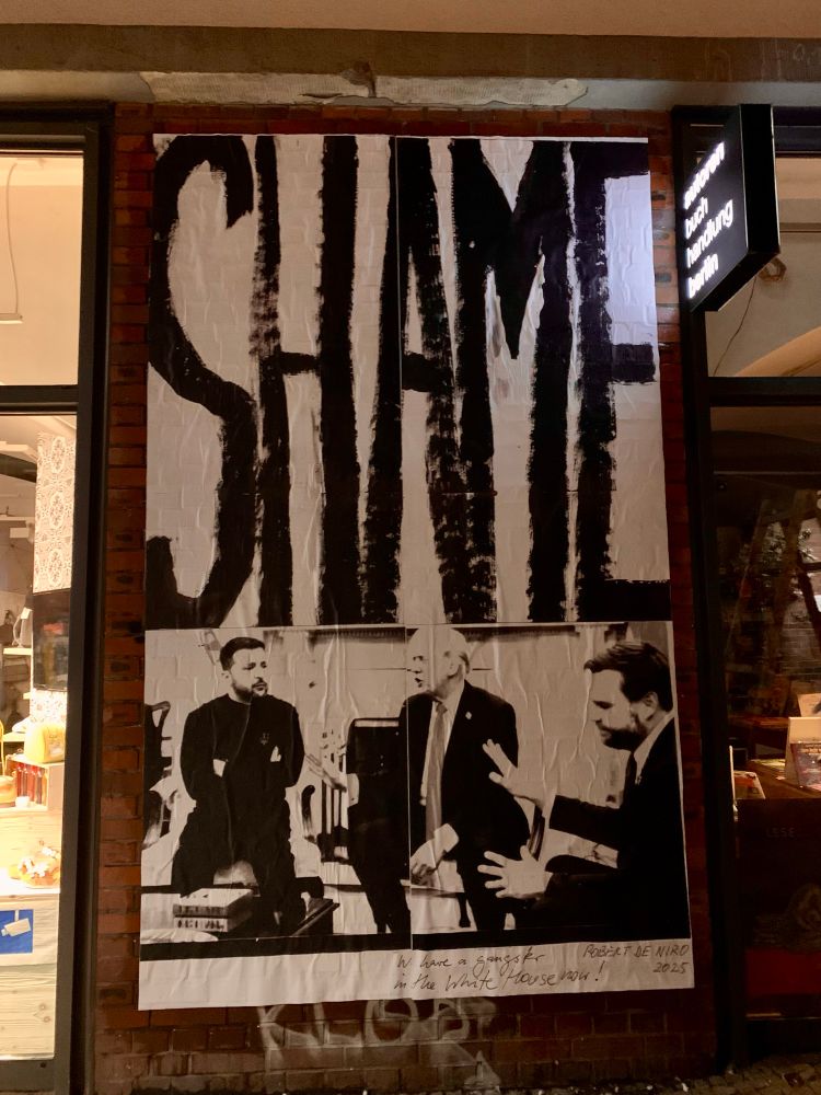 Ein etwa drei Meter hohes Wandplakat: iIn großen, steilen, schwarzen Buchstaben das Wort Shame, darunter eine Szene mit Selensky und Trump im Weißen Haus. Unter dem Bild ein Zitat von Robert der Niro: We have a gangster in the White House