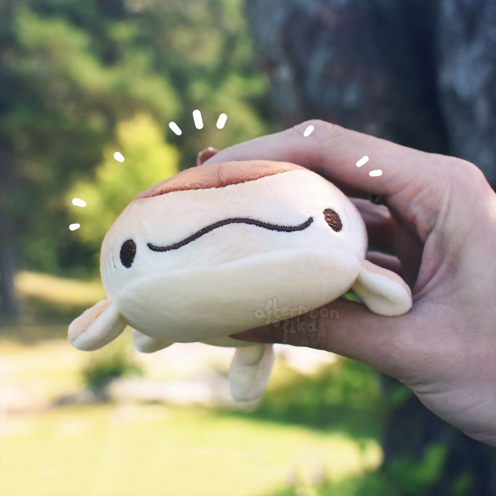 A photo of a chocolate doughnut whale shark keychain plush being squished gently from above and below by a hand. The plush is seen from the front, showing its goofy little smile. So sweet!