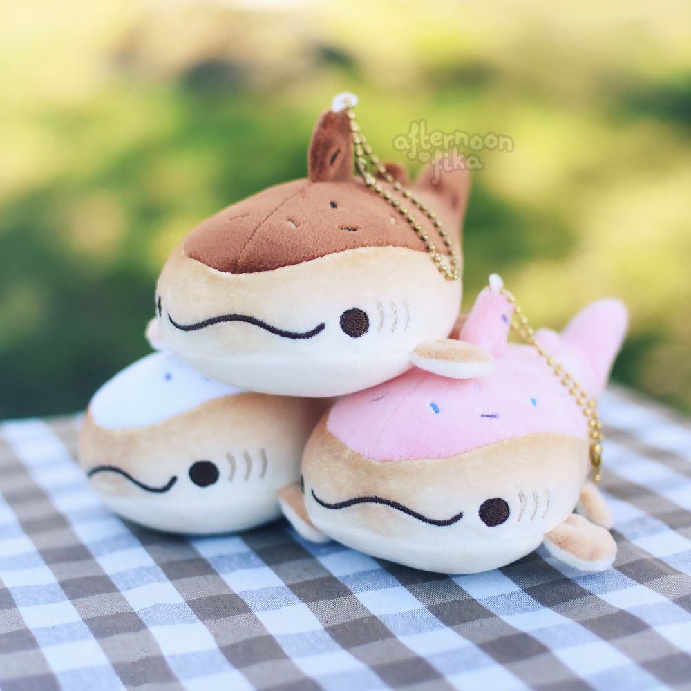 Three doughnut whale shark keychain plushies stacked in a triangle formation on top of a brown and white picnic blanket. The two bottom plushies are strawberry and sugar flavoured, with colourful sprinkles on them. The top plushie is chocolate flavoured, with sprinkles being various shades of brown. Their dorsal fins have the keychain itself attached to them, and it is gold. What a delightful assortment of sweets!