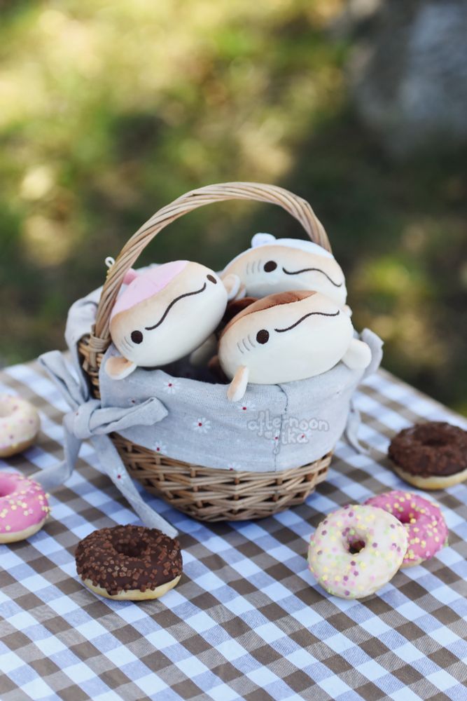Three doughnut whale shark keychain plushies neatly placed in a small brown basket, their little faces peeking out. The basket is on top of a brown and white picnic blanket, and around it, small doughnuts have been placed. The flavours of the plushies, much like their pastry counterparts, are chocolate, sugar and strawberry. Better keep them in the shade, so their frosting won’t melt!