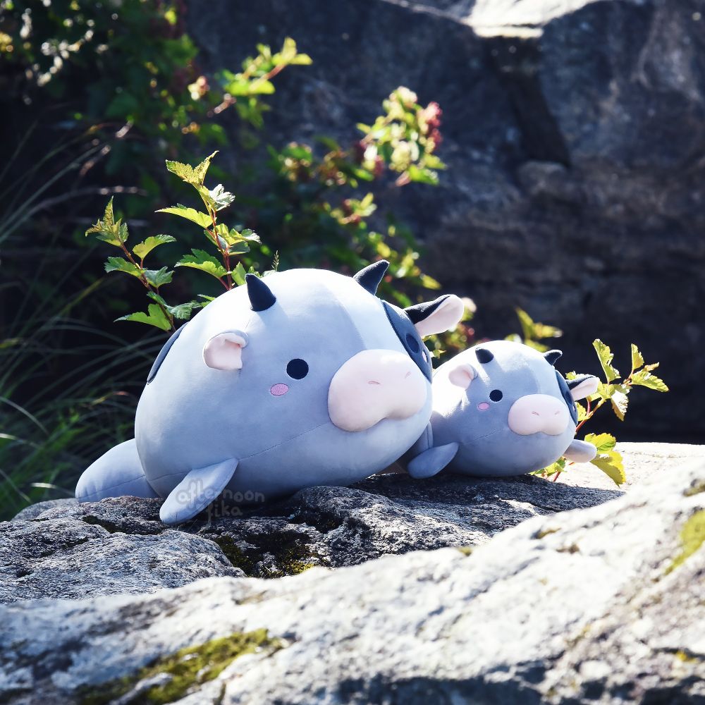 A photo of two sea cow, or manatee, plushies, next to each other and on top of a rock under the sun. The sea cow on the left is larger and round, around 50cm in length, with pink cheeks and a ver rotund body. Her daughter, to her right, has the same proportions, but is around 30cm long instead. They are both enjoying a little sunbathing on a fine summer morning. How lovely!