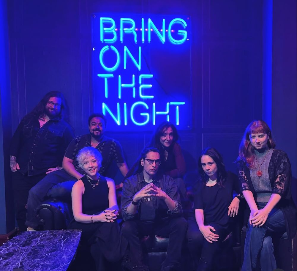 Johnny Chiodini, Jasper William Cartwright, Belinda Cornish, Mark Meer, Samantha Béart, Paula Deming and Josephine sit on a couch posing in front of a neon sign that reads “Bring on the night,” tingeing the whole photo with a cool blue tint!